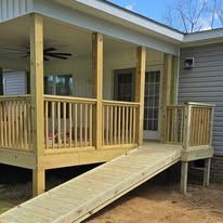 Wooden porch with ramp and railing attached to a gray house, under a covered roof.