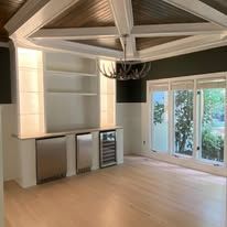 Modern empty room with vaulted ceiling, light wood floors, built-in shelving, and glass doors opening to greenery
