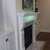 White fireplace mantel with black insert in a living room, beside white cabinets and a rug.