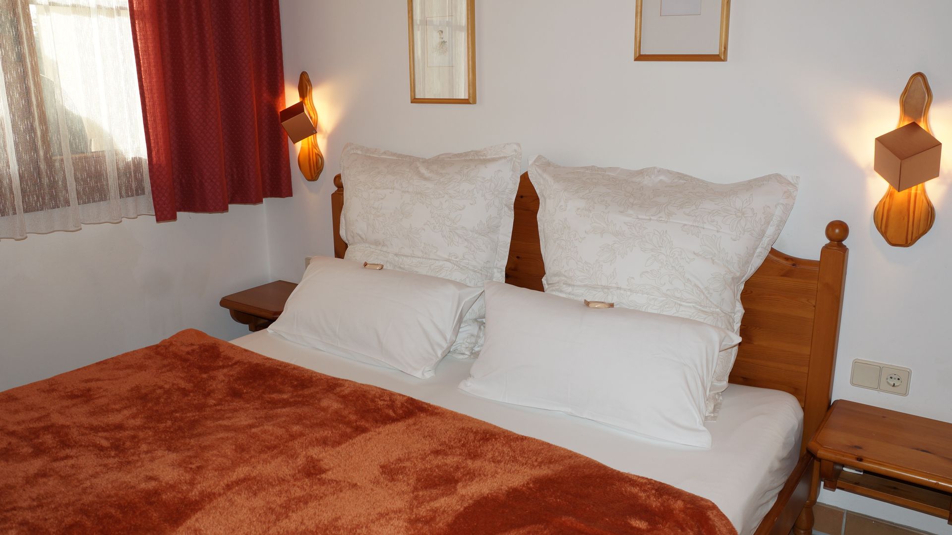 Gemütliches Schlafzimmer mit ordentlich gemachtem Bett, weißen Kissen, roter Decke und warmen Wandlampen.