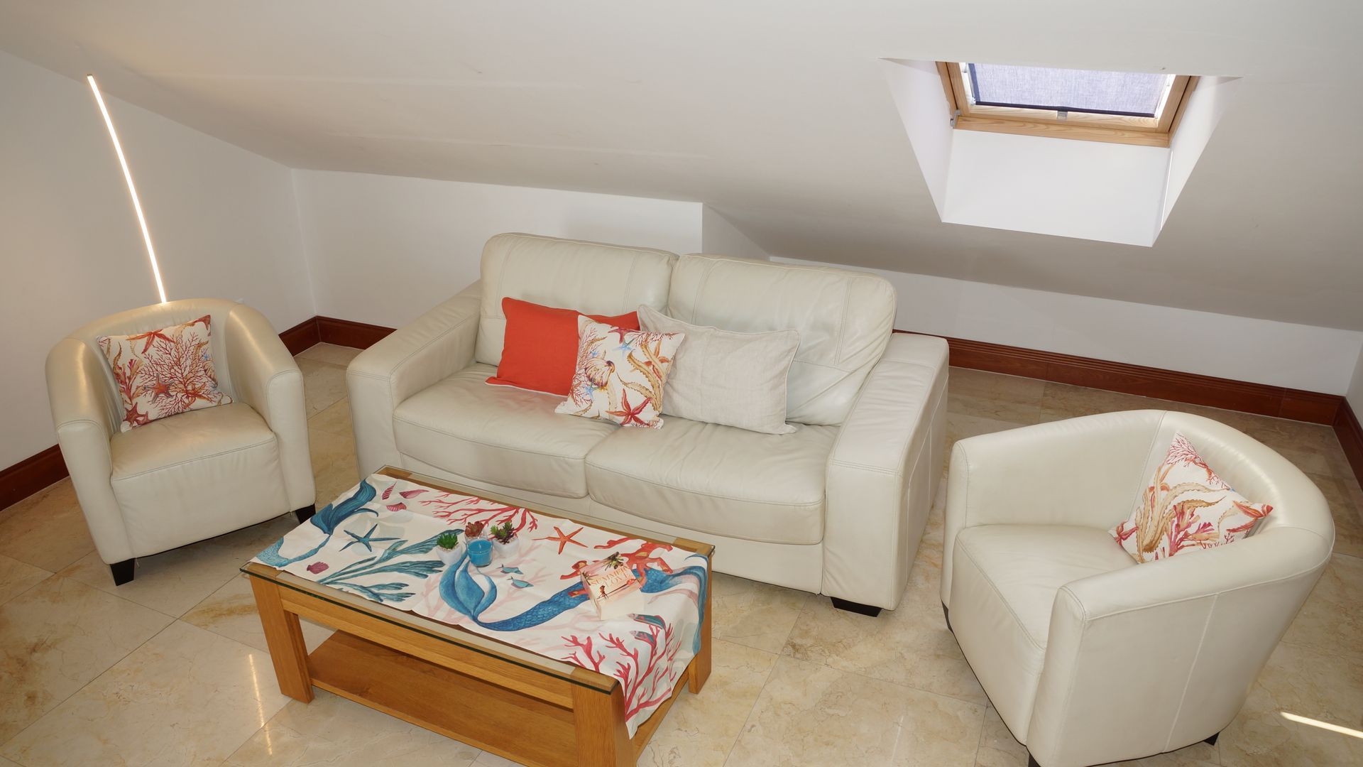 Weißes Wohnzimmer mit zwei Sesseln, einem Sofa und einem farbenfrohen Couchtisch unter Dachfenstern.