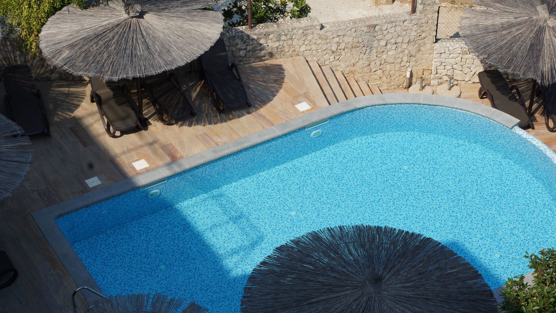 Ein blauer Swimmingpool neben strohgedeckten Sonnenschirmen und Liegestühlen in einem sonnigen Innenhof eines Ferienresorts