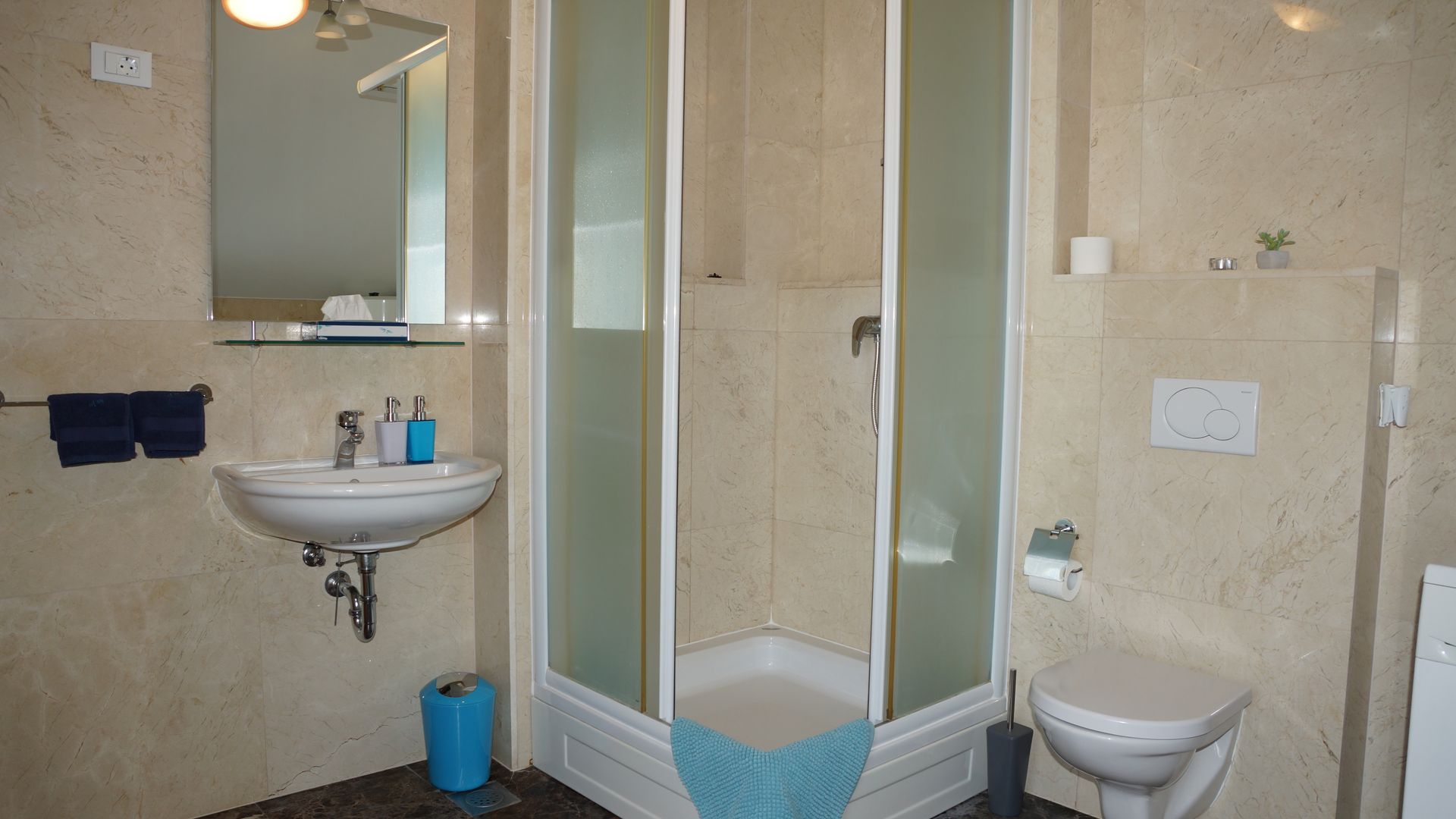 Kleines beigefarbenes Badezimmer mit Eckdusche, Waschbecken, Toilette, Spiegel und blauen Akzenten
