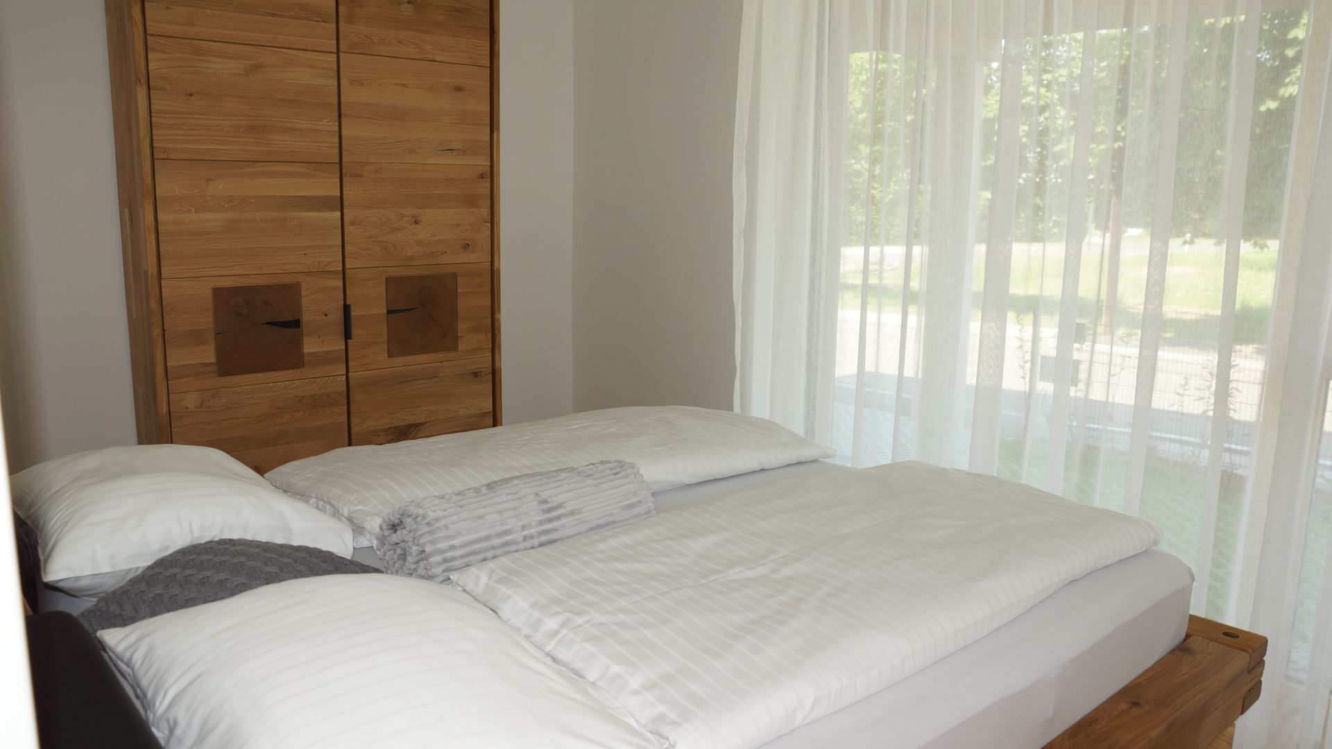 Ein ordentlich gemachtes Bett mit weißer Bettwäsche neben einem Holzschrank in einem sonnendurchfluteten Schlafzimmer