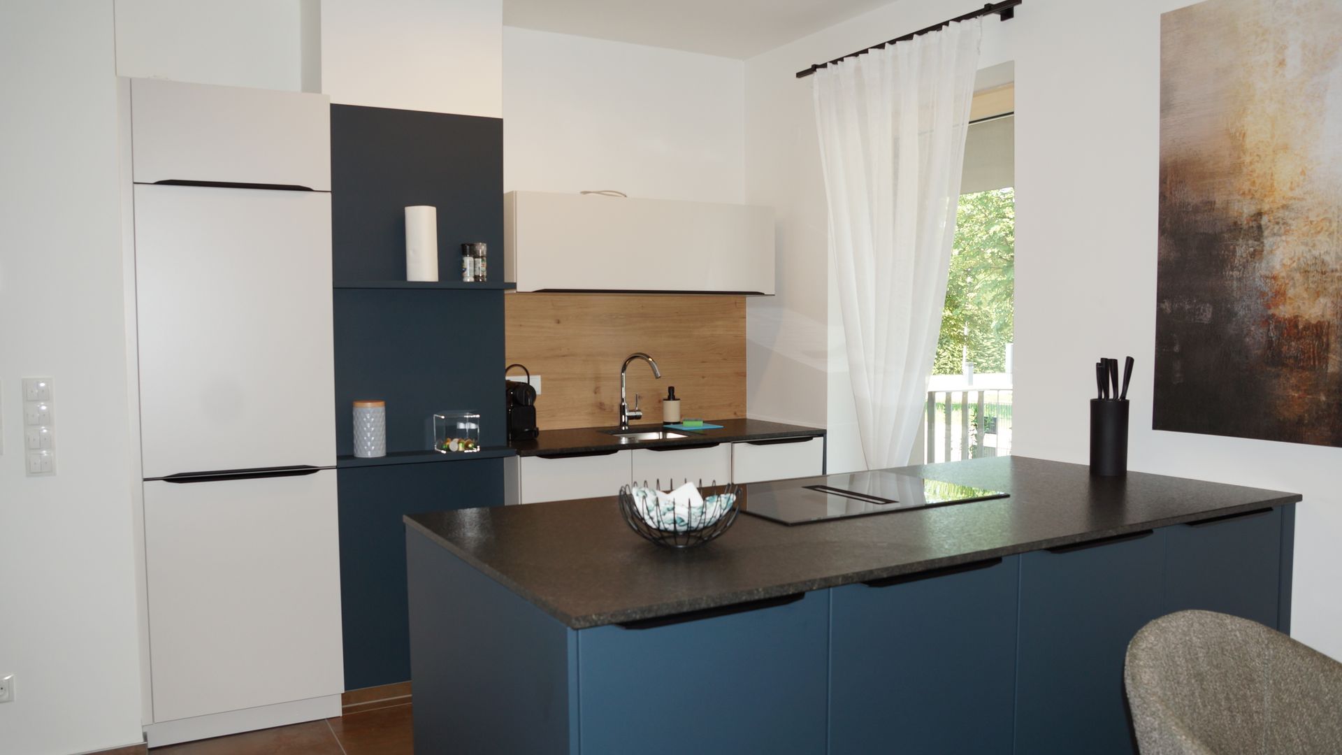 Moderne Küche mit dunkler Kochinsel, weißen Schränken und einem Fenster mit transparenten Vorhängen.