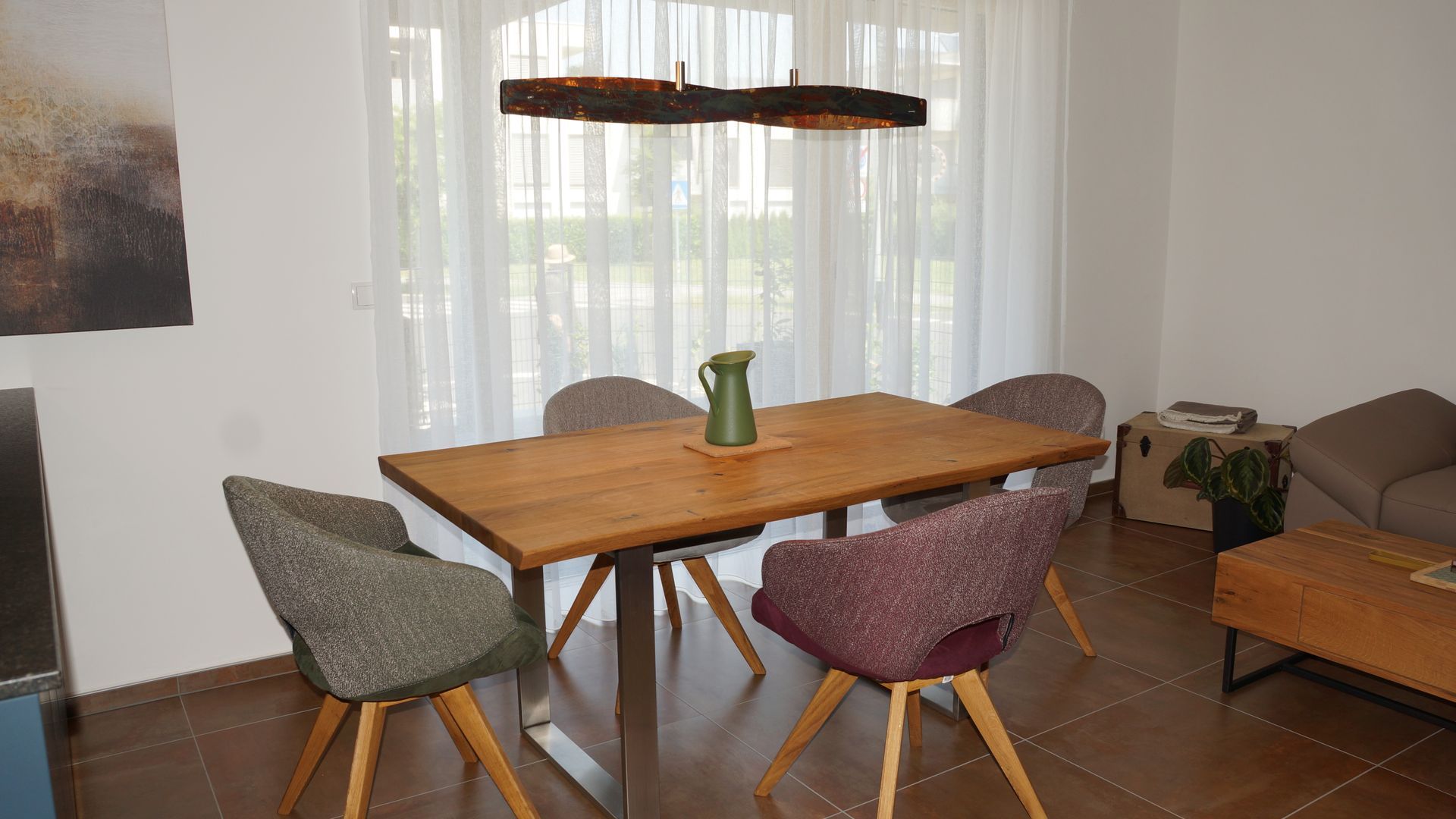 Modernes Esszimmer mit Holztisch, unterschiedlichen Stühlen und einer Vase vor transparenten Vorhängen.
