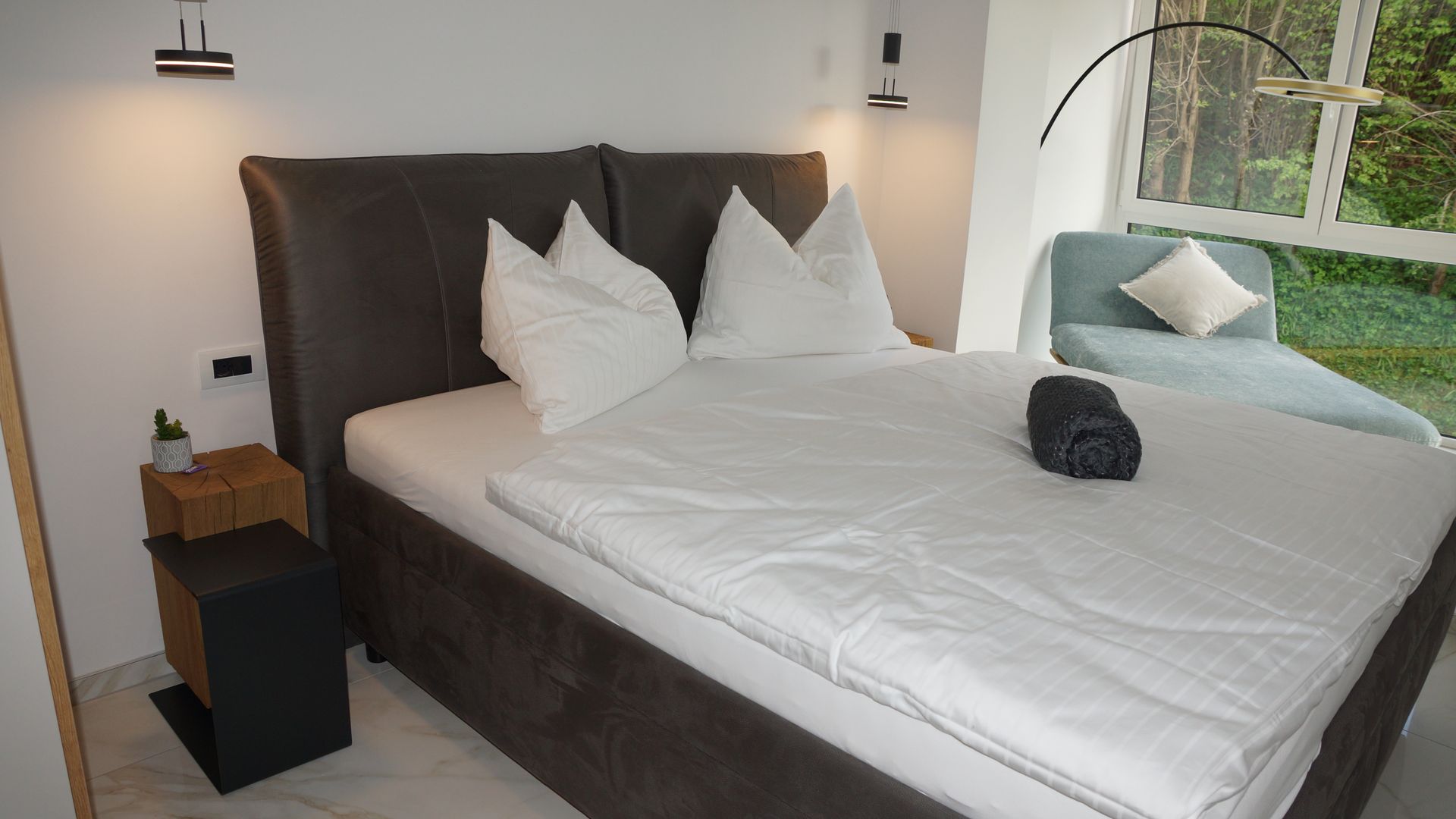 Gemütliches Schlafzimmer mit ordentlich gemachtem Bett, weißer Bettwäsche, dunklem Kopfteil und großer Fensterbank.