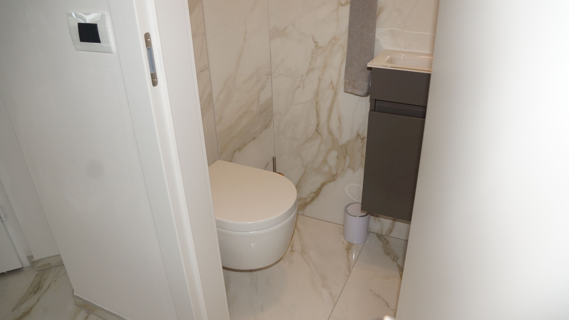 Kleines Badezimmer mit Toilette, beigefarbenen Wänden in Marmoroptik und einem teilweise sichtbaren dunklen Waschtisch.