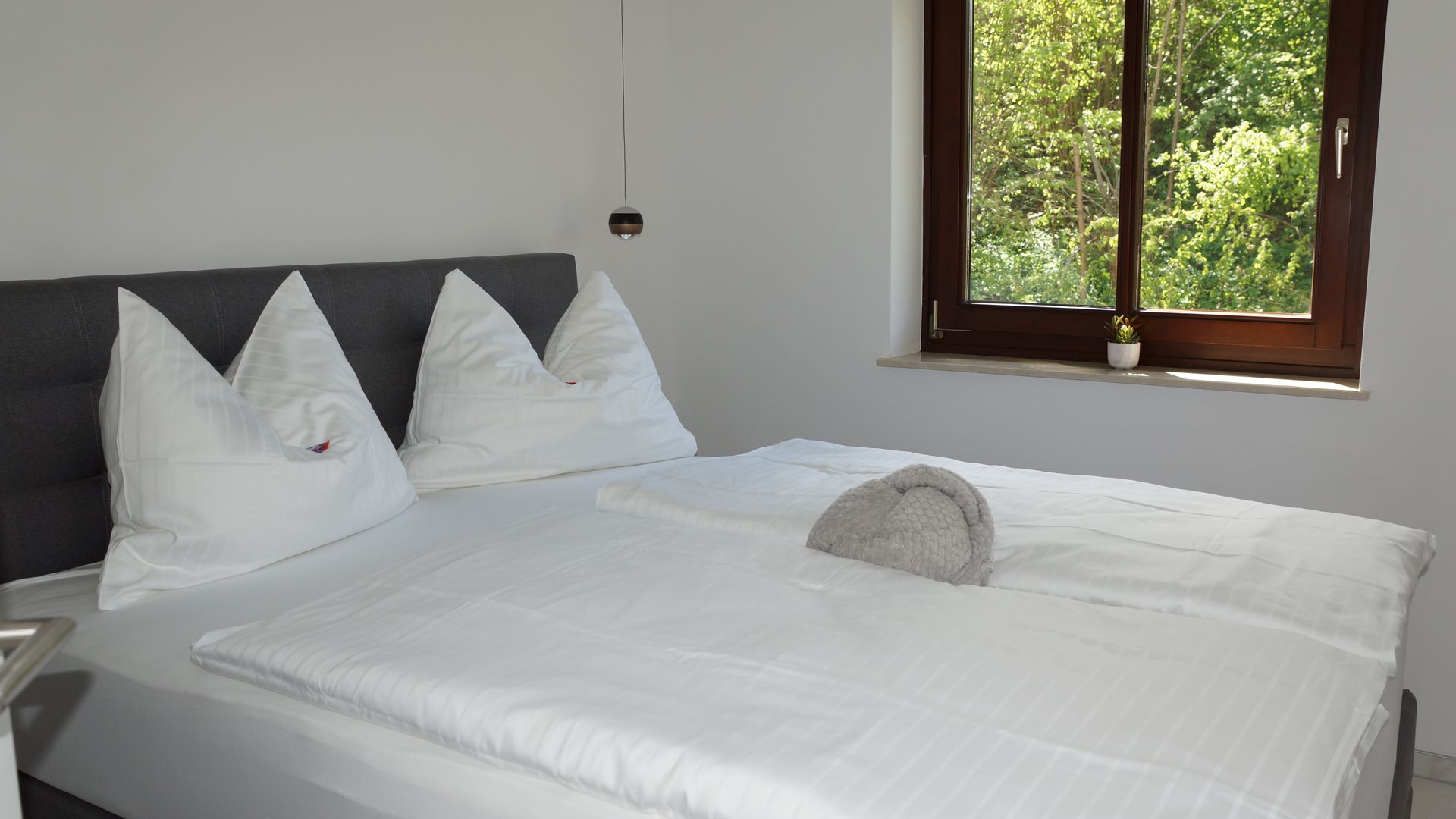 Ein ordentlich gemachtes weißes Bett in einem hellen Zimmer neben einem Fenster mit Blick ins Grüne.