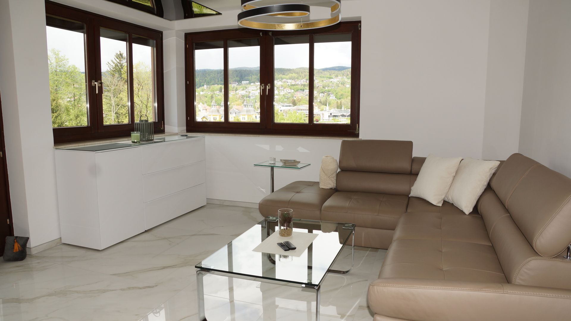 Helles Wohnzimmer mit beigefarbenem Ecksofa, Glas-Couchtisch und großen Fenstern mit Blick ins Grüne