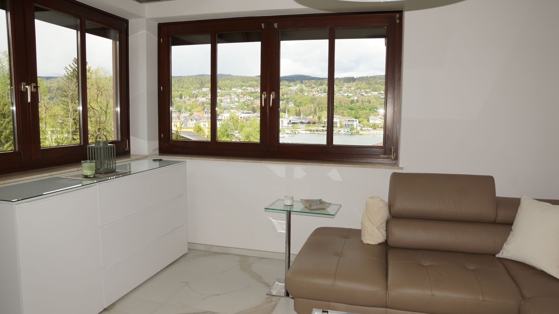 Modernes Wohnzimmer mit beigefarbenem Sofa, weißen Schränken und großen Fenstern mit Blick ins Grüne