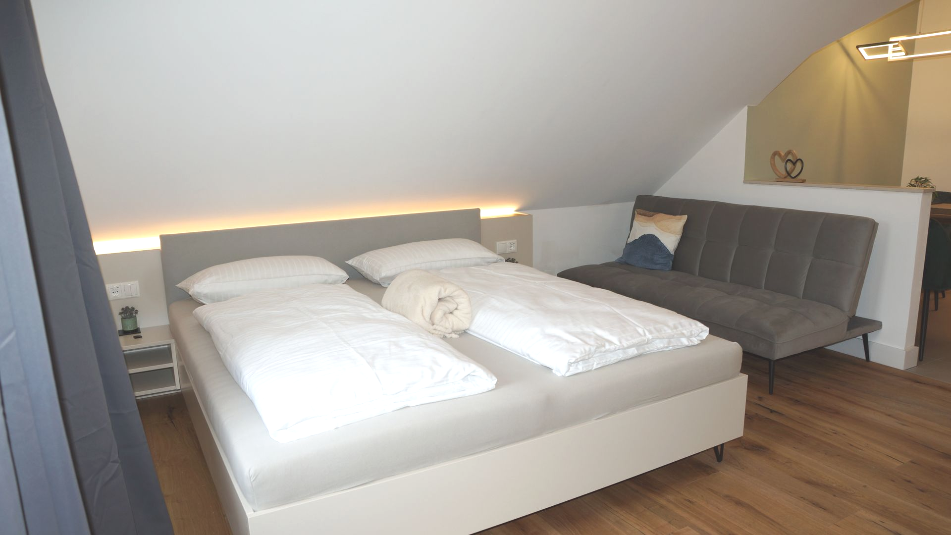 Dachgeschoss-Schlafzimmer mit zwei weißen Betten, einem grauen Sofa und warmer Akzentbeleuchtung unter einer Dachschräge