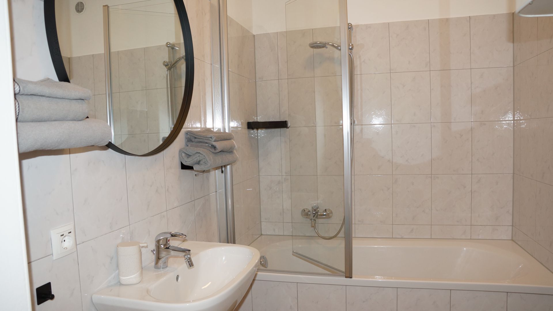 Modernes Badezimmer mit Waschbecken, Wandspiegel, Glasdusche und Badewanne mit beigefarbenen Fliesen.