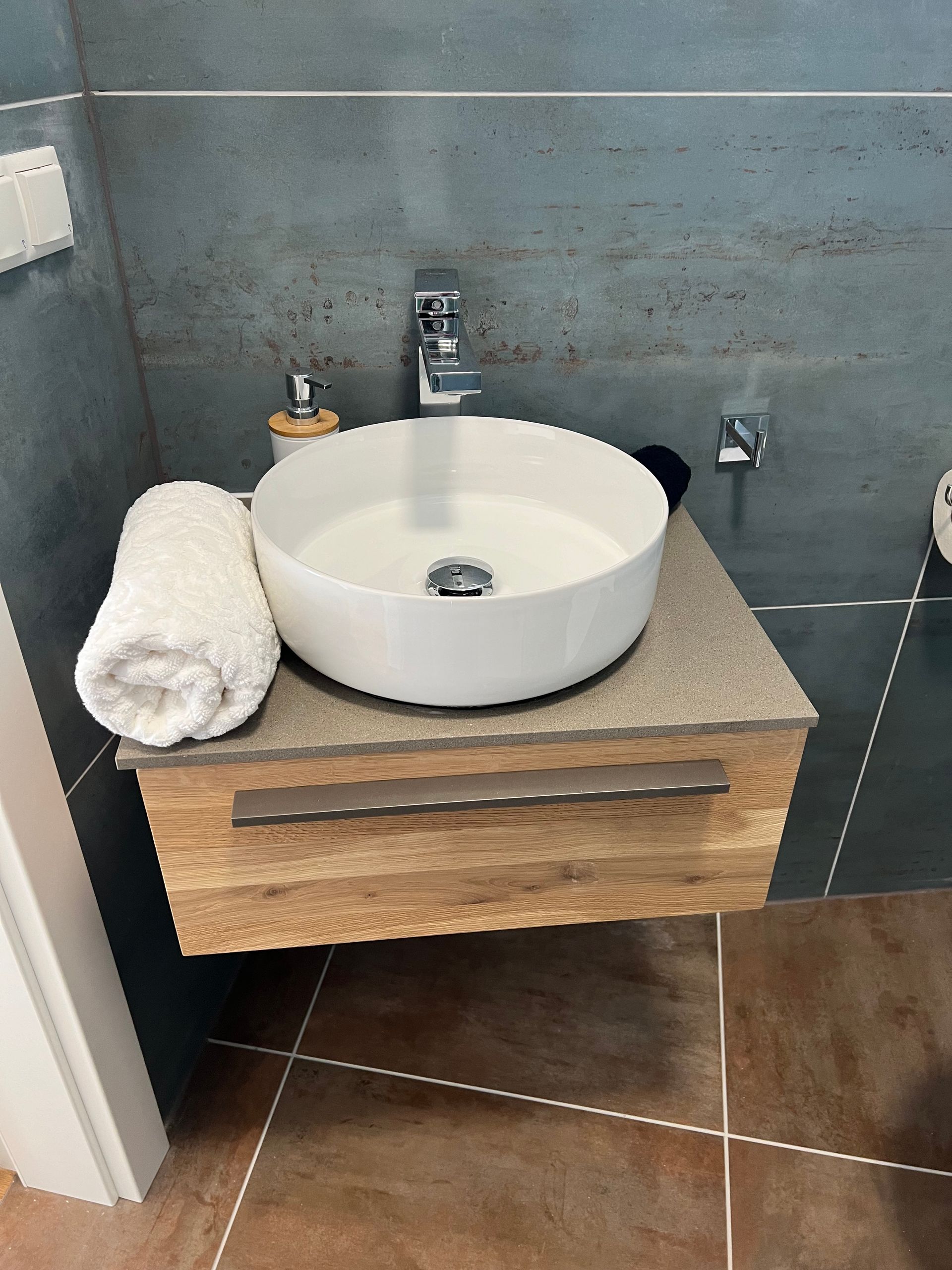Moderner Badezimmerwaschtisch mit rundem, weißem Waschbecken, Holzschrank, grauer Fliesenwand und gefaltetem Handtuchhalter