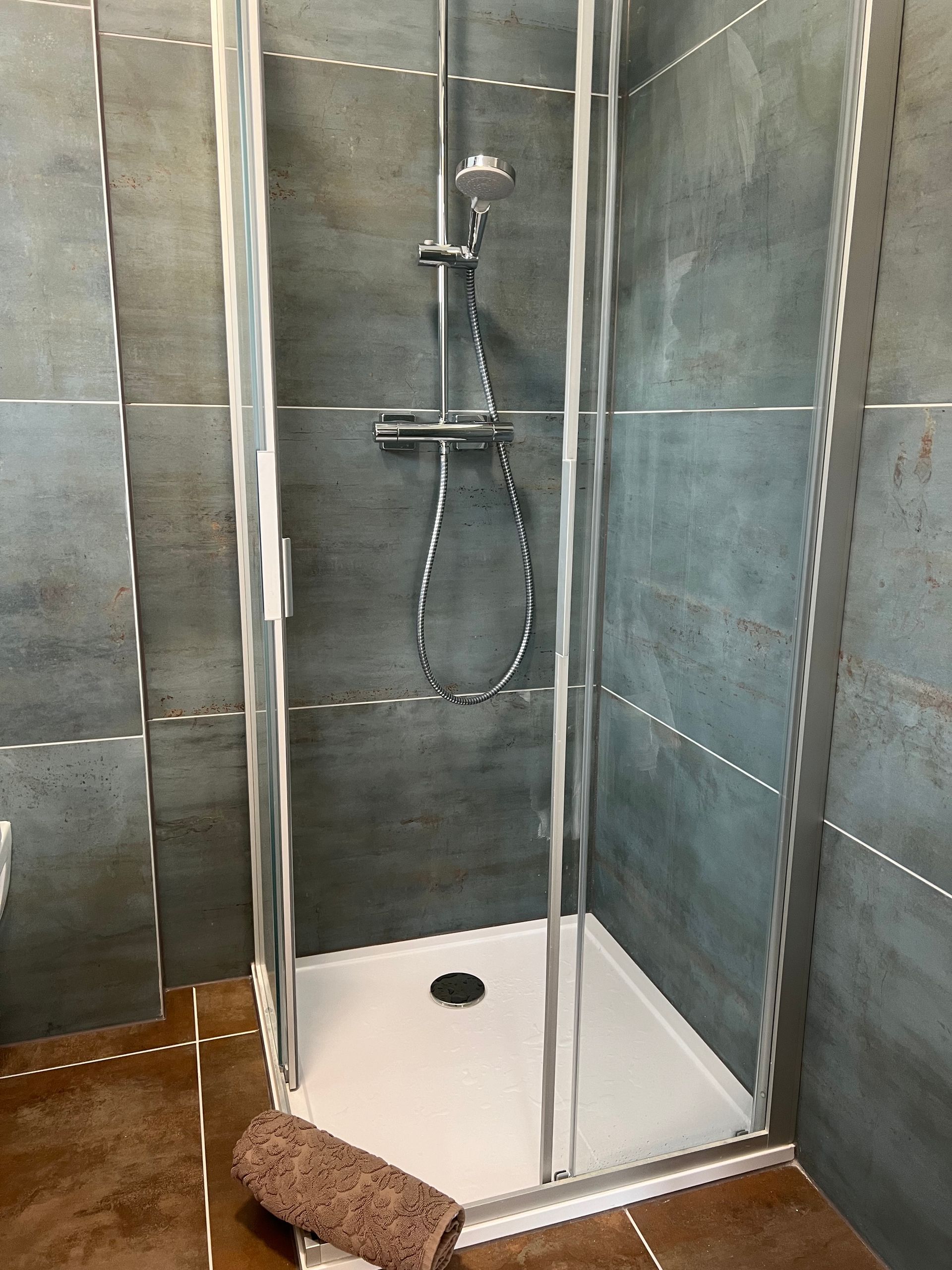 Begehbare Dusche mit Glastüren, Handbrause, grauen Fliesenwänden und einer braunen Badematte.
