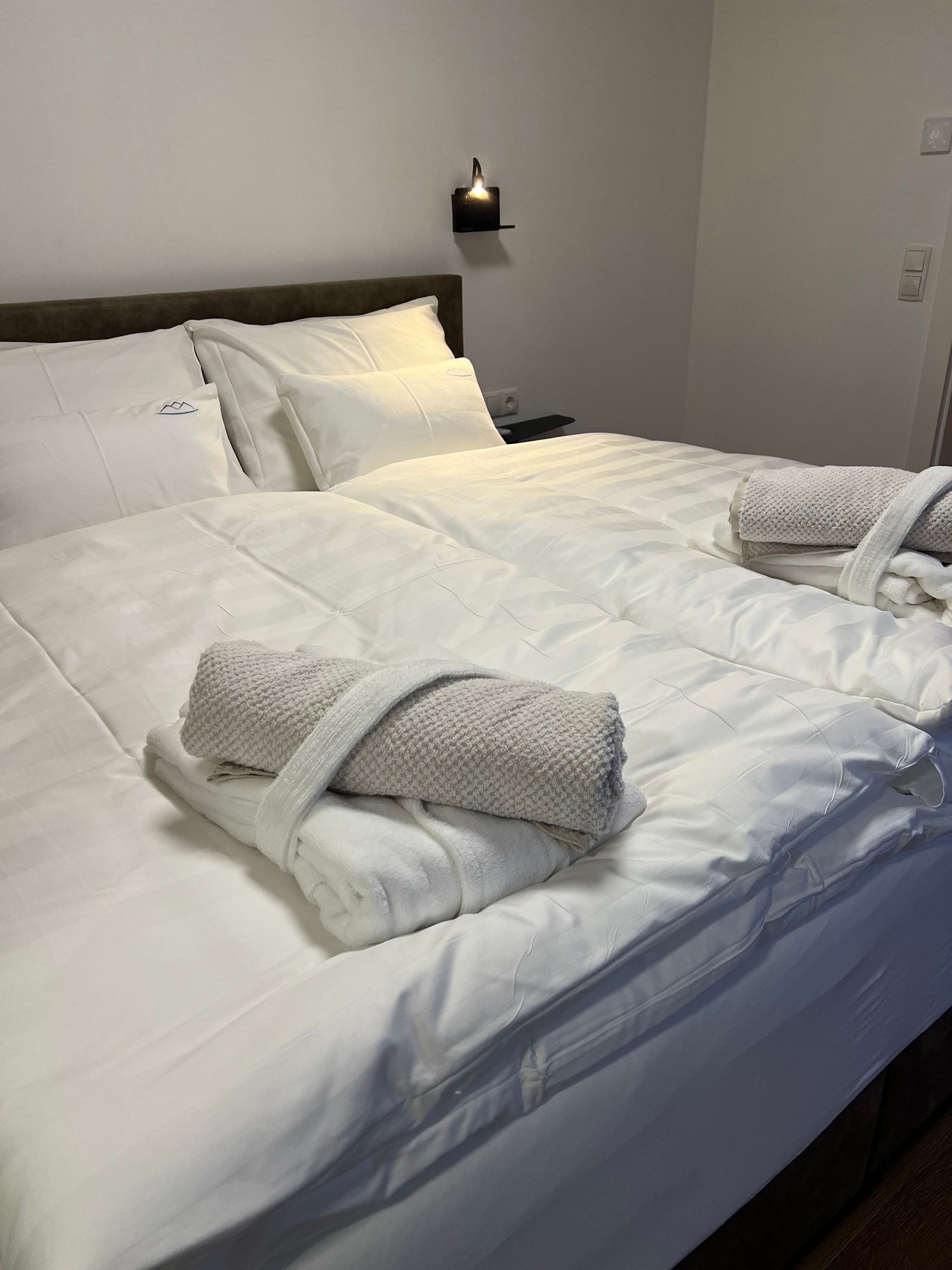 Ein ordentlich gemachtes weißes Bett in einem modernen Schlafzimmer mit gefalteten Handtüchern und einer Leselampe.