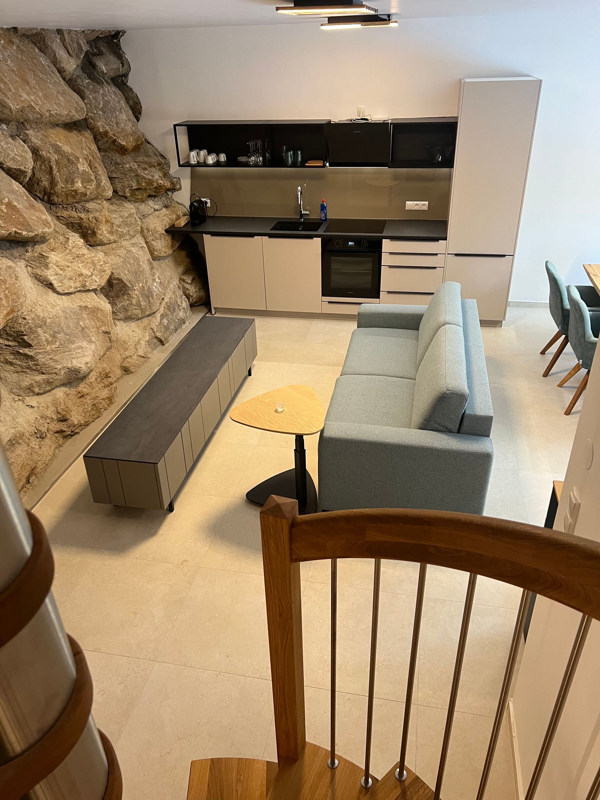 Modernes, offenes Wohnzimmer mit grauem Sofa, Küche, Steinwand und kleinem Tisch.