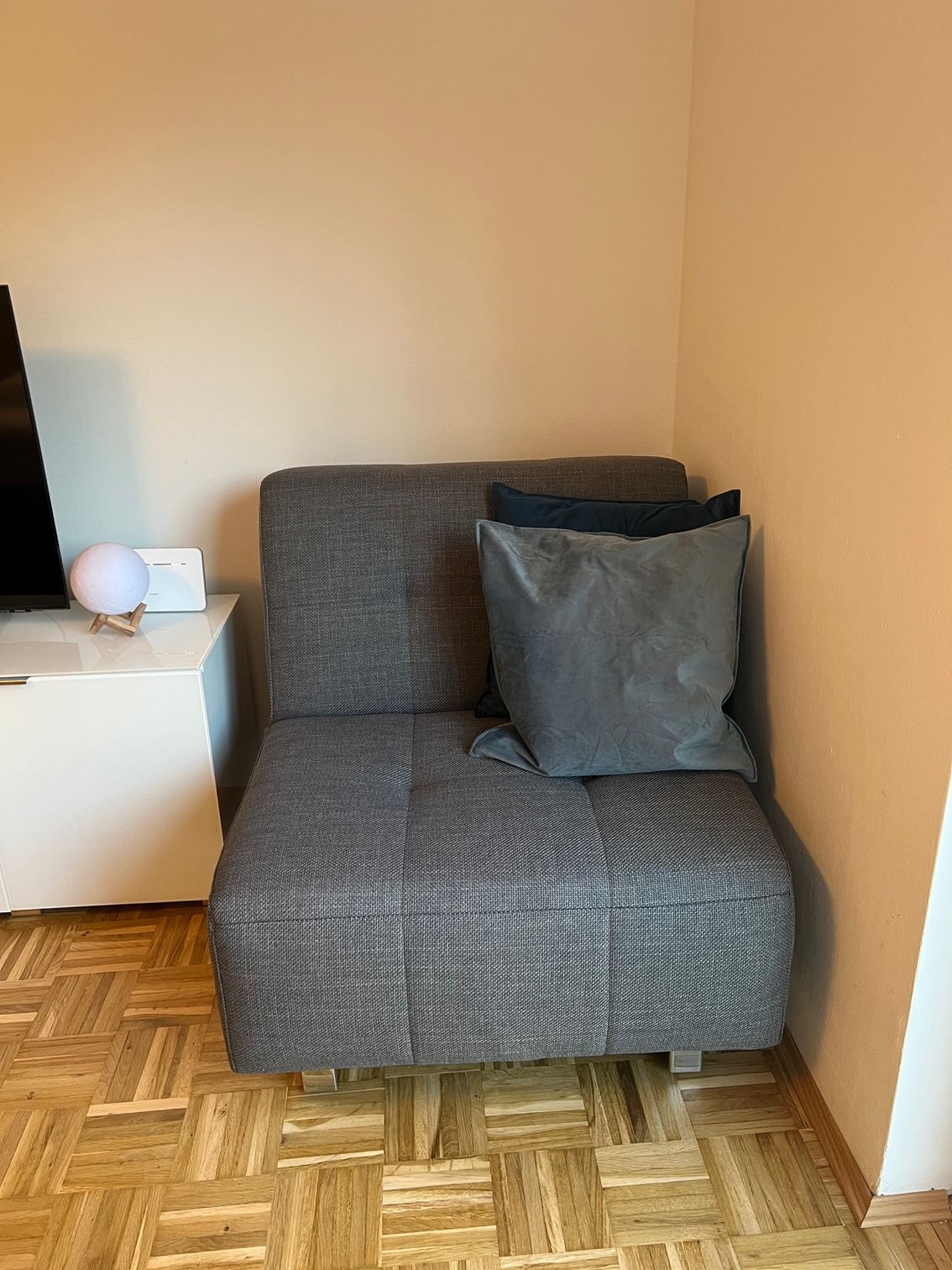 Ein grauer Stuhl in einer Ecke eines beigen Zimmers neben einem weißen Schrank auf Parkettboden.