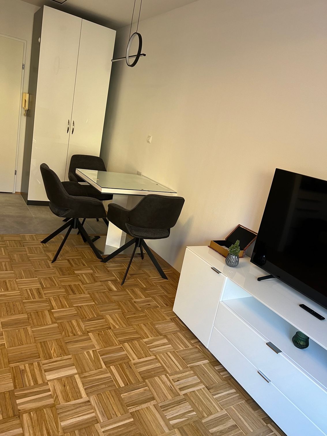 Kleine Ess-/Arbeitsecke mit schwarzem Stuhl, weißem Tisch, Kleiderschrank und Fernseher auf einer weißen Kommode.
