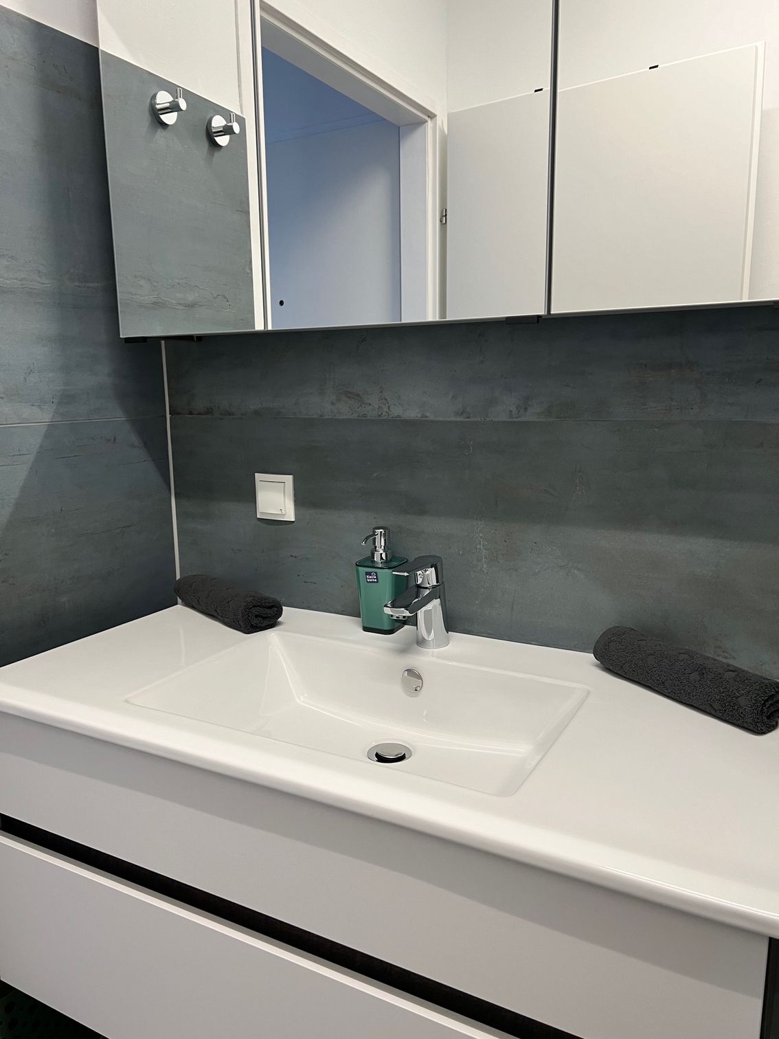 Moderner Badezimmerwaschtisch mit weißem Waschbecken, grau gefliester Wand, Spiegelschrank und türkisfarbenem Seifenspender.