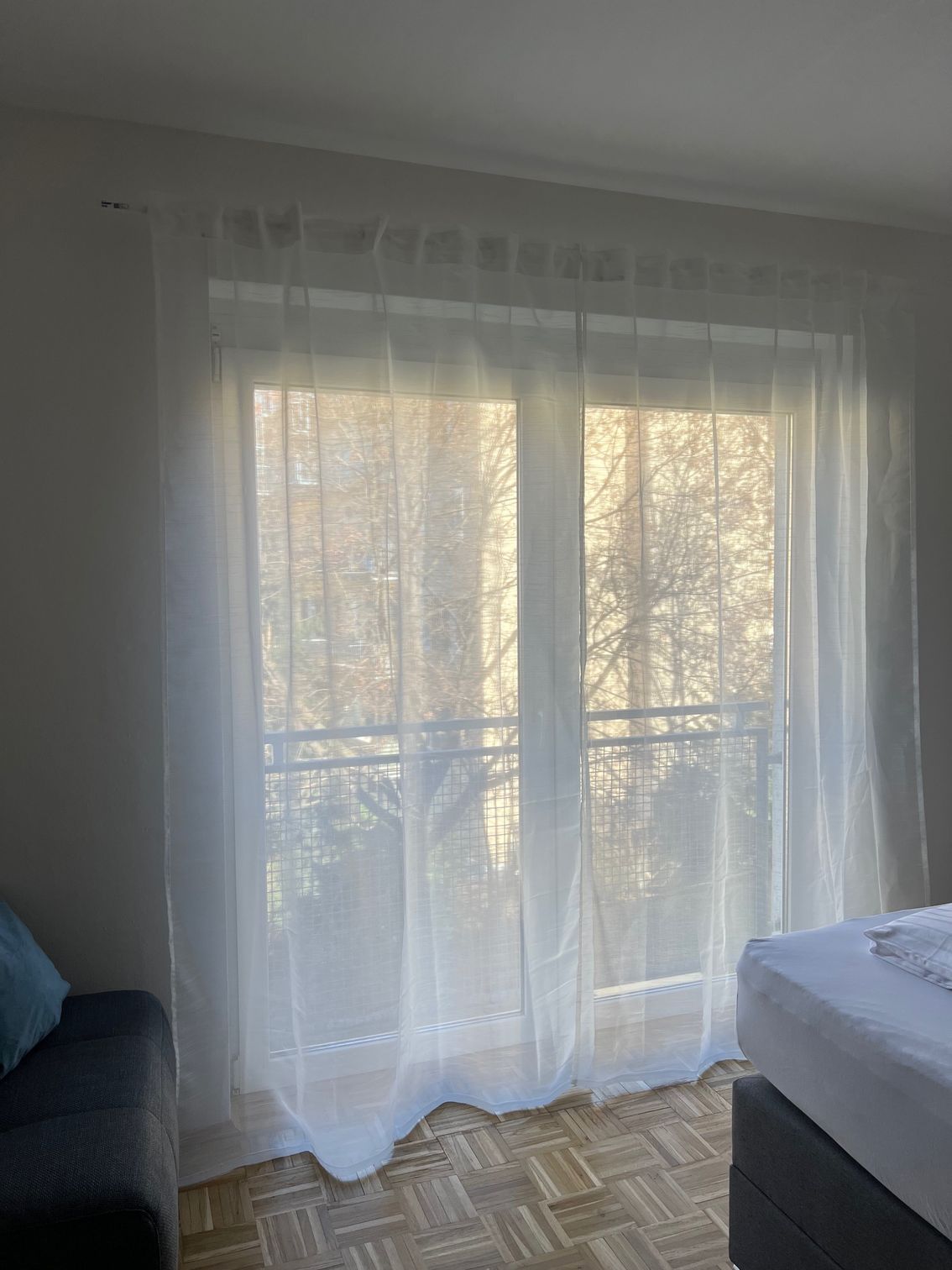 Transparente Vorhänge vor einer sonnendurchfluteten Balkontür in einem Schlafzimmer, rechts ein Bett und links ein Sofa.