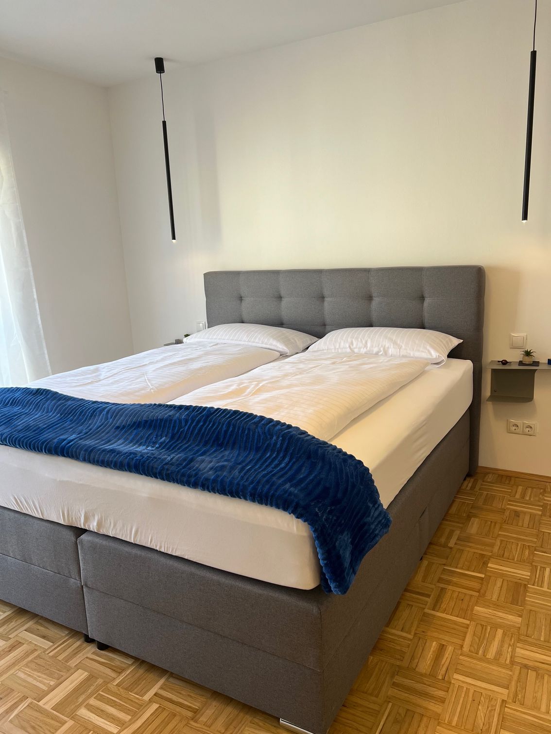 Ein ordentlich gemachtes graues Bett mit weißer Bettwäsche und einer blauen Tagesdecke in einem Schlafzimmer