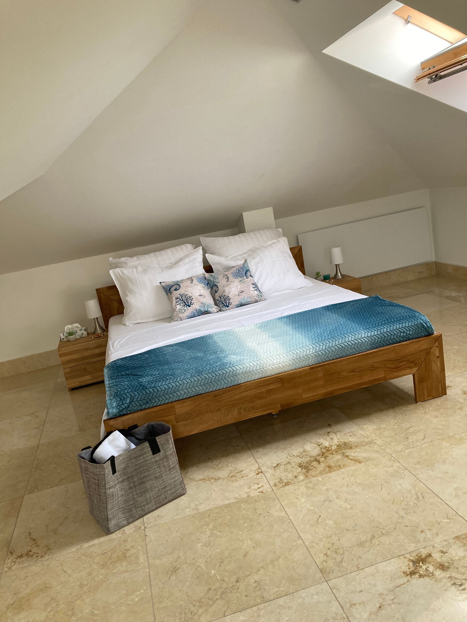 Minimalistisches Dachgeschoss-Schlafzimmer mit Holzbett, weißer Bettwäsche, blauer Tagesdecke und Dachfenster.