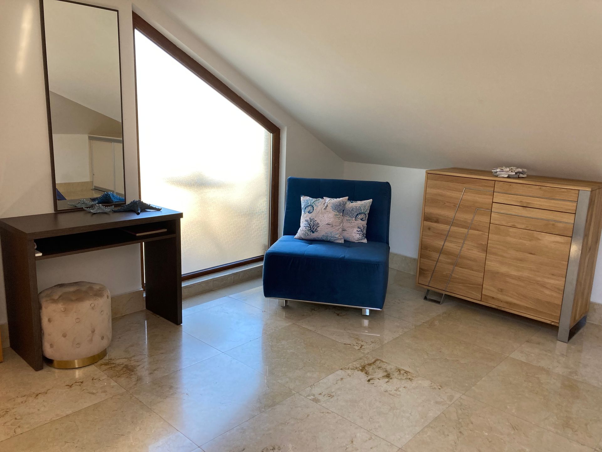 Dachzimmer mit blauem Sessel, Schminktisch und Holzkommode an einem schrägen Fenster.