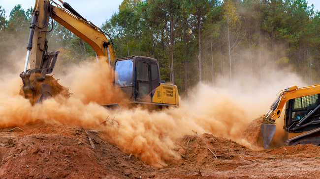 Excavator digging in red dirt, kicking up a large cloud of dust in a wooded construction site