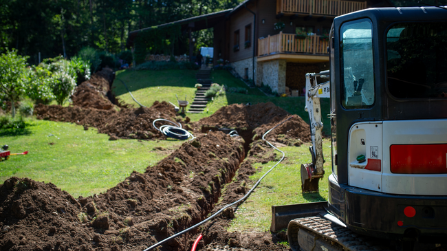 Backhoe digging a long trench beside a house in a grassy yard