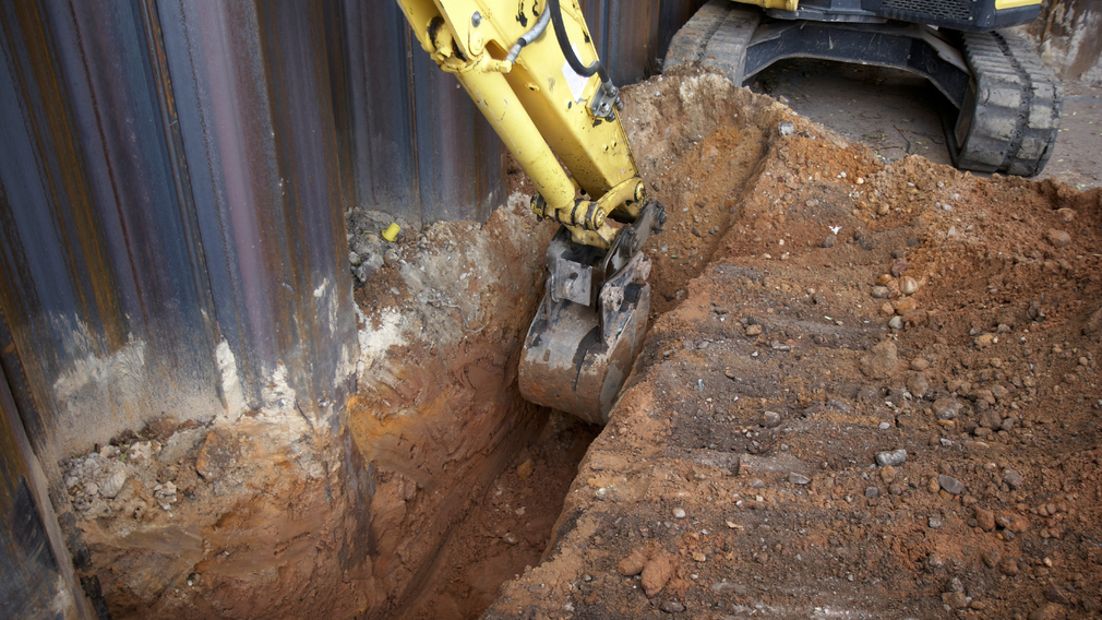 Yellow excavator digging a trench beside a corrugated metal wall, moving wet brown soil