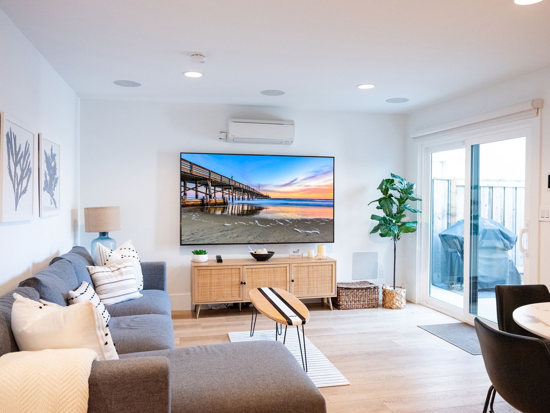 Bright living room with gray sofa, wall-mounted TV, wooden console, plants, and sliding glass doors to a patio