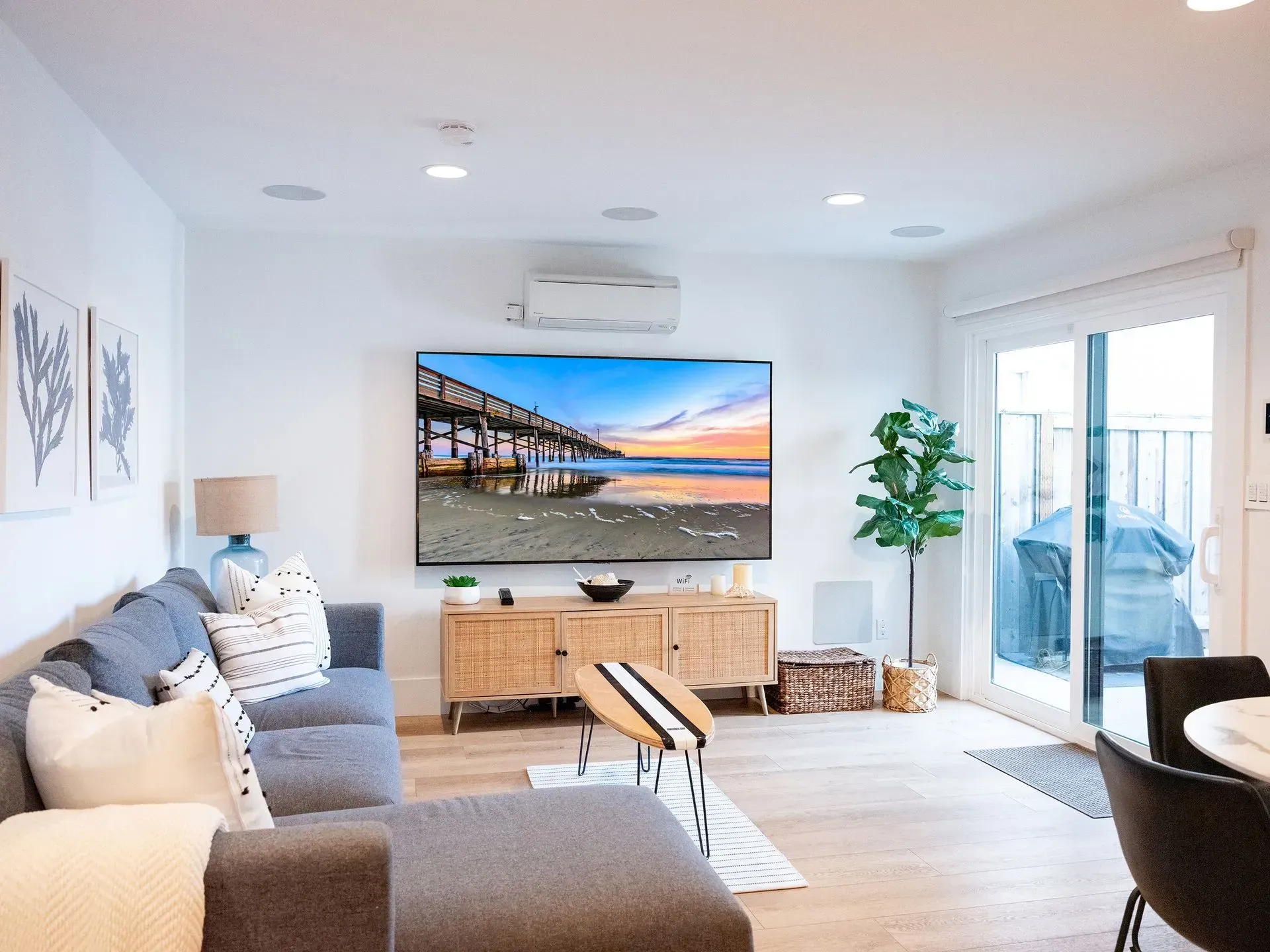 Bright living room with gray sofa, wall-mounted TV, wooden console, plants, and sliding glass doors to a patio