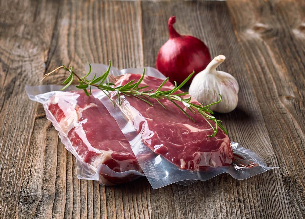 Um bife selado a vácuo está sobre uma mesa de madeira ao lado de alho e cebola.