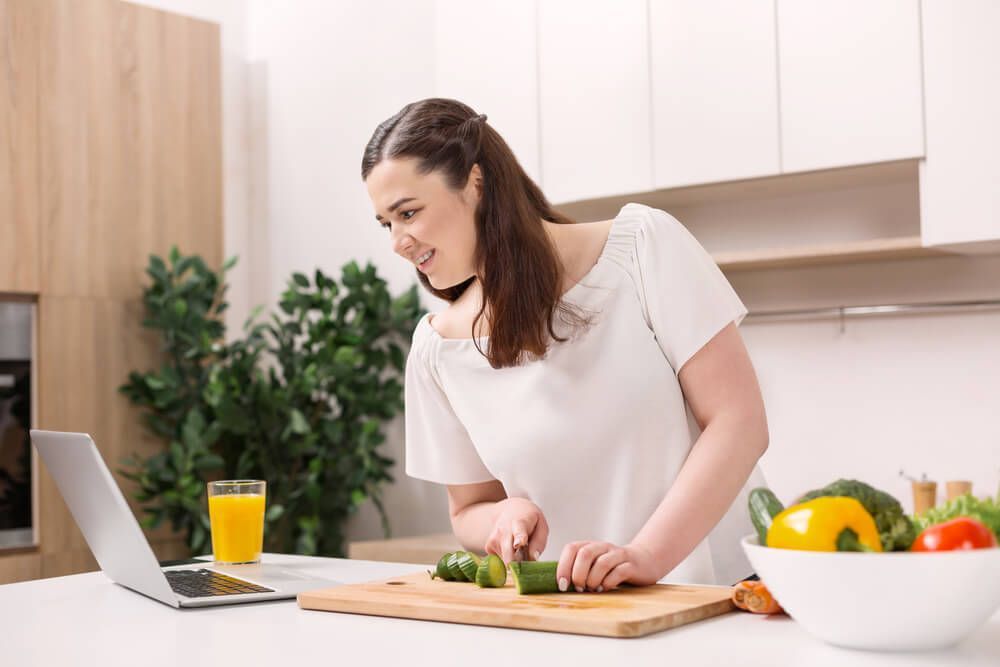 Uma mulher está cortando vegetais na cozinha enquanto olha para um laptop.