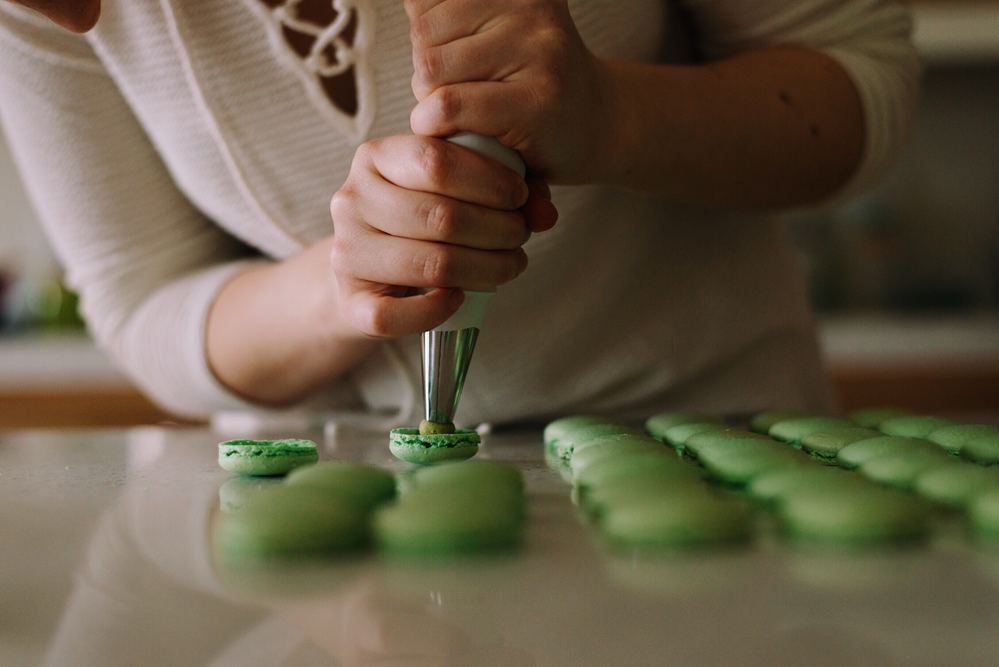 Uma mulher está decorando macarons verdes com um saco de confeitar.