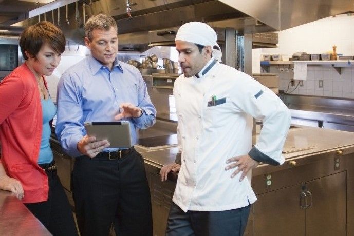 Um homem está olhando para um tablet enquanto está ao lado de um chef na cozinha.
