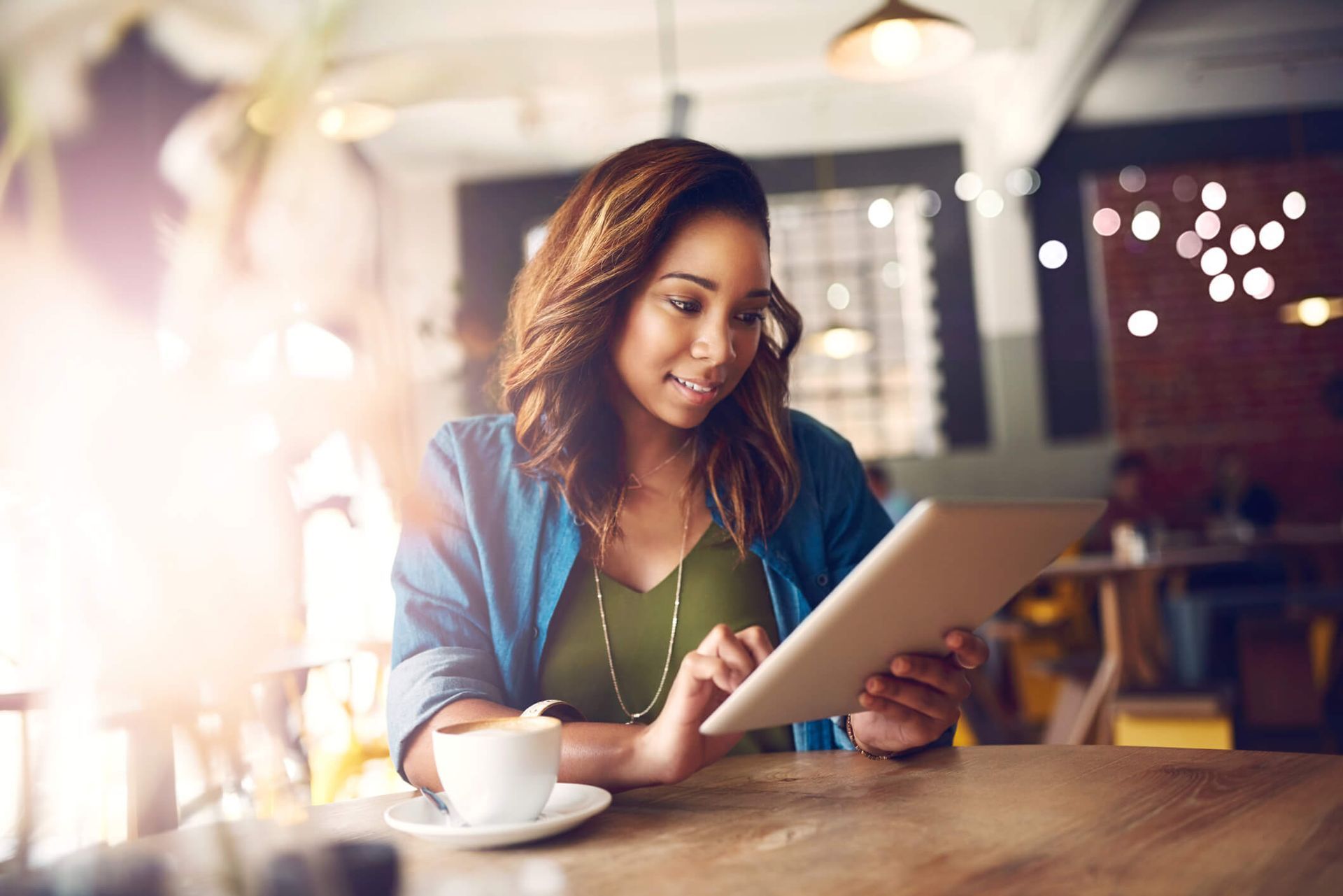 Uma mulher está sentada à mesa com uma xícara de café e um tablet.