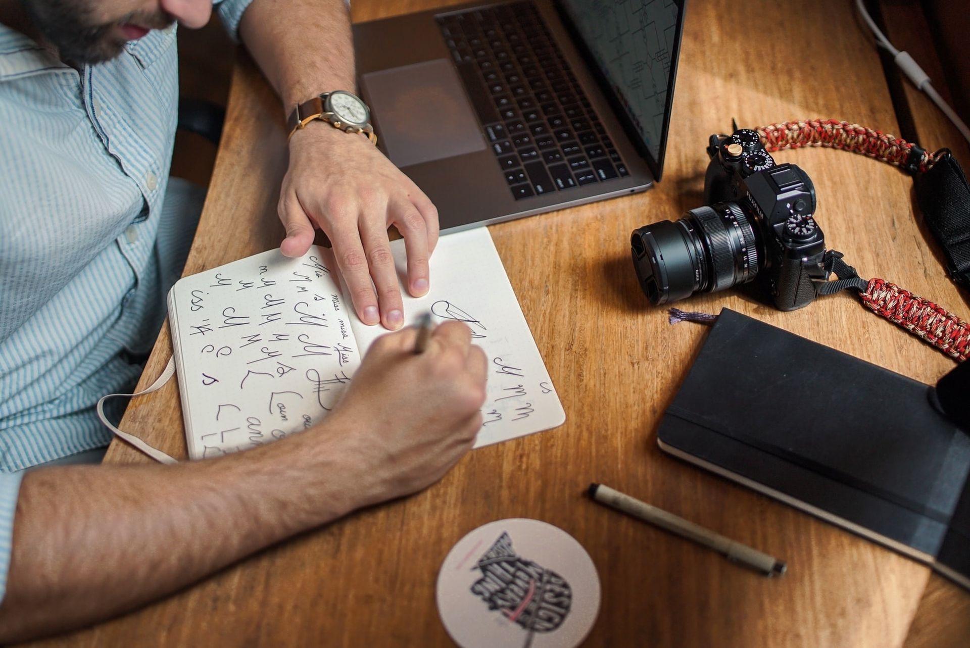 Um homem está sentado à mesa escrevendo em um caderno ao lado de um laptop e uma câmera.