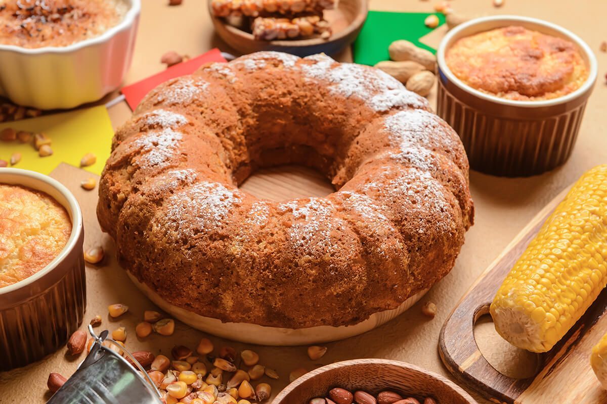 Um bolo bundt com açúcar de confeiteiro por cima está sobre uma mesa ao lado de espigas de milho.