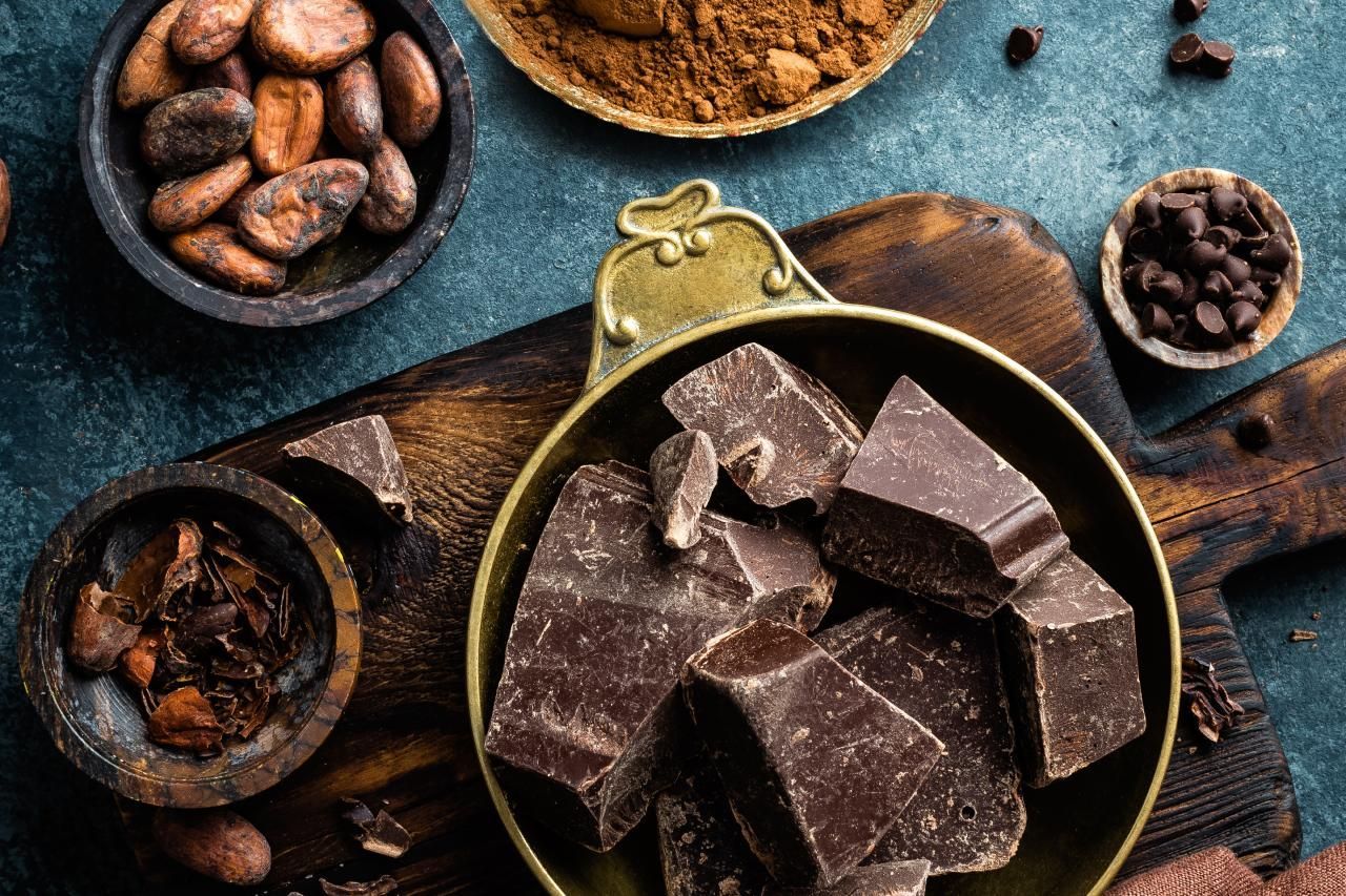Uma tigela de chocolate está sobre uma tábua de corte de madeira ao lado de uma tigela de grãos de cacau.