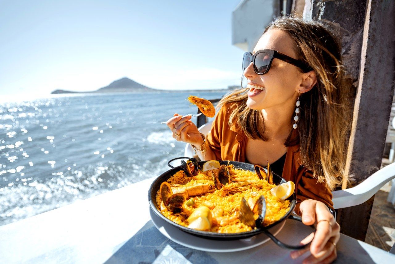 Uma mulher está sentada à mesa comendo paella à beira-mar.