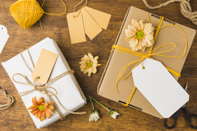 Duas caixas embrulhadas em papel branco e amarradas com barbante e flores sobre uma mesa de madeira.