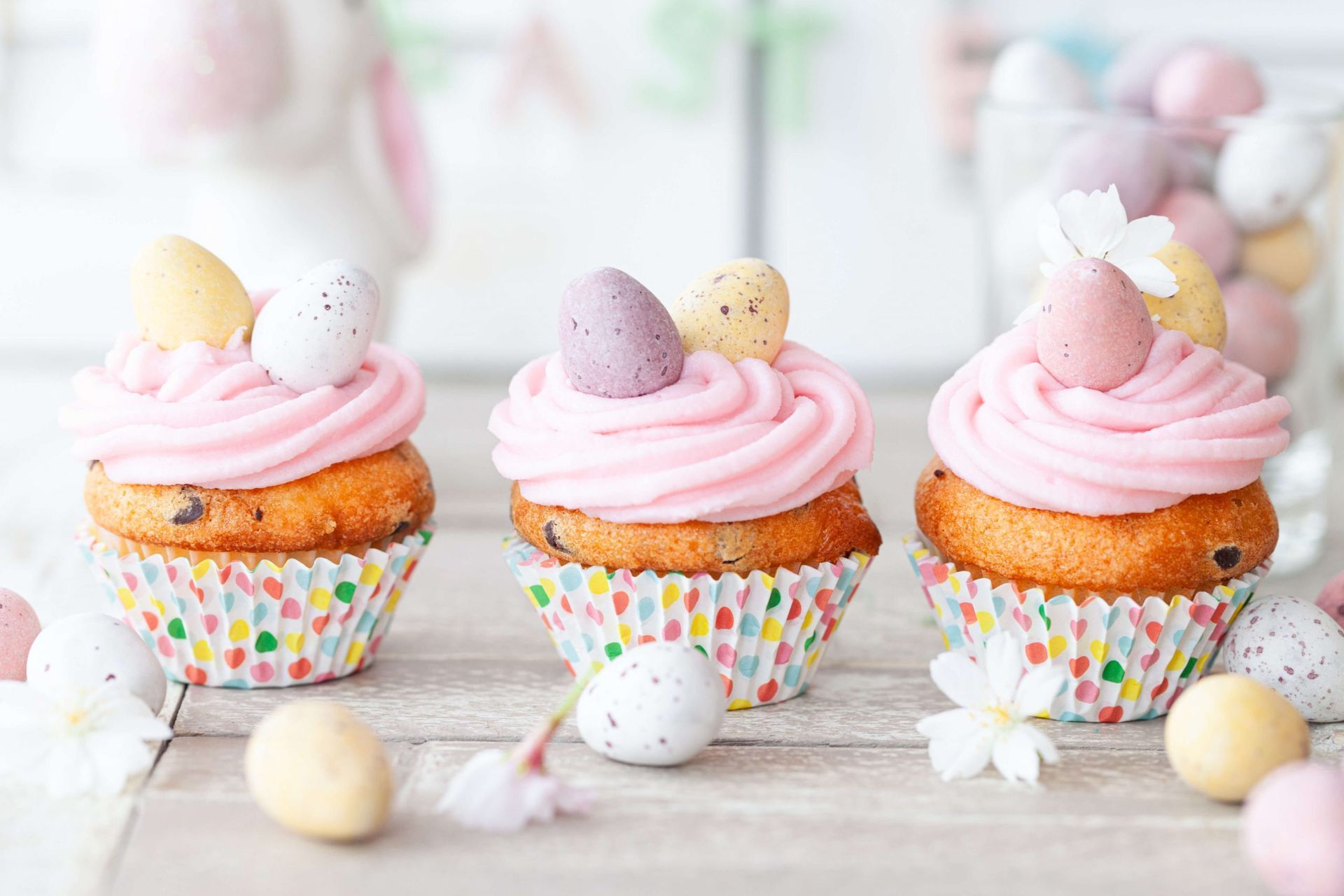 Três cupcakes com cobertura rosa e ovos de páscoa no topo estão sobre uma mesa de madeira.