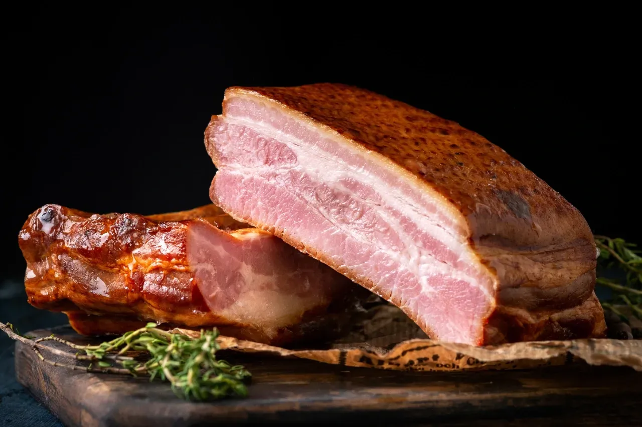 Peça de bacon defumado sobre tábua de madeira, acompanhada de ervas frescas, com fundo escuro