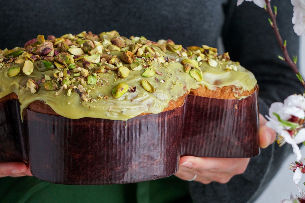 Uma pessoa com uma colomba pascal com cobertura de pistaches e flores ao fundo.