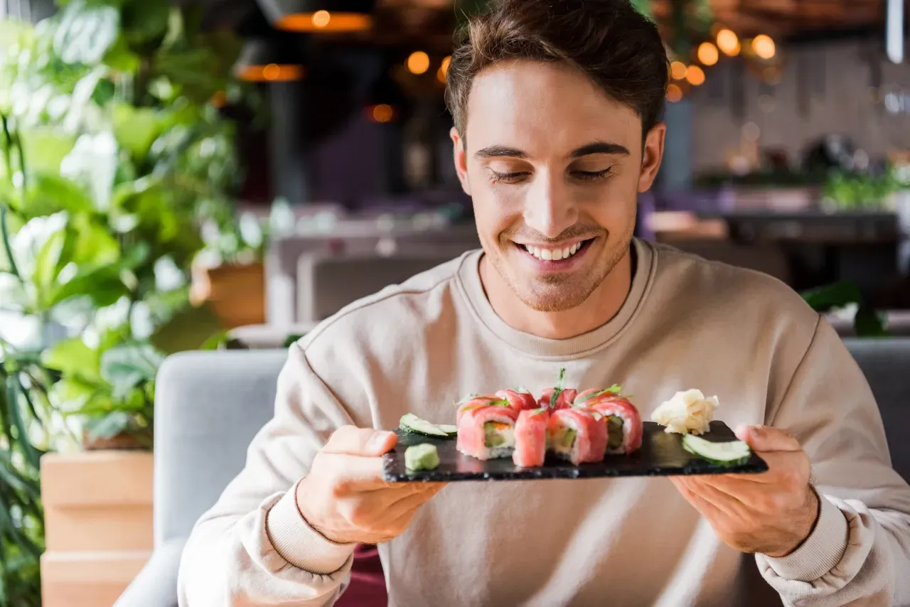 Homem sorrindo enquanto come sushi sozinho em um restaurante, segurando uma pequena porção individual de sushi em uma bandeja.