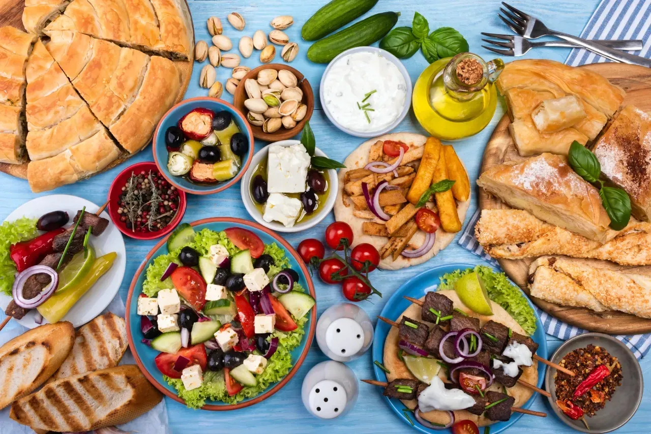 Mesa com pratos da comida grega, incluindo salada grega, souvlaki, pão pita, queijo feta, iogurte grego, azeitonas, spanakopita e azeite de oliva.