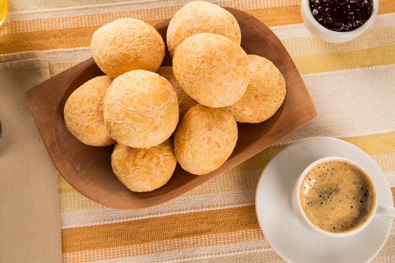 Tigela de madeira com pãezinhos de queijo dourados ao lado de uma xícara de café com geleia.