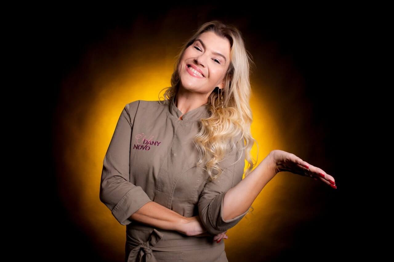 Uma mulher em uniforme de chef está sorrindo e estendendo a mão.